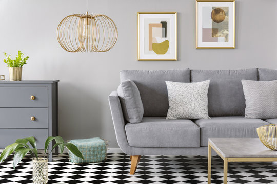 Gold lamp in grey living room interior with poster above grey sofa on checkered floor. Real photo
