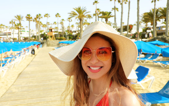 Smiling young woman with straw hat and sunglasses on the beach looking at the camera. Happy beautiful girl with deck chairs, umbrellas and palm trees, typical tropical scenery at Canary Islands, Spain