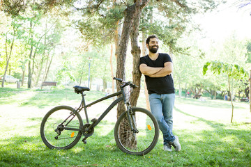 Obraz premium Smiling man leaning on a tree in the park with his bicycle next to him