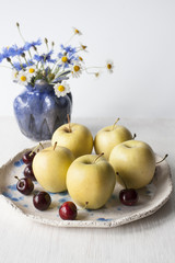 Yellow apples and juicy cherries lie on a plate of clay. Nearby is a pigeon vase with field flowers. Composition on a white wooden background.