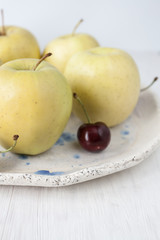 One juicy red cherry and a few yellow tasty apples in a clay plate on a white wooden background. The view is close..