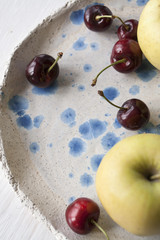 A few juicy red cherries and yellow apples in a clay plate on a white wooden background. Top view.