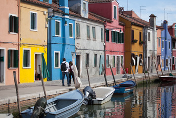 Fototapeta na wymiar 2 people walking - Houses of Burano and reflection in the water. Waterways with traditional boats and colorful facade. Venice - Italy