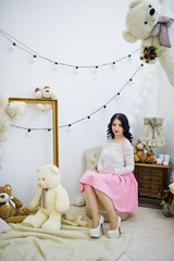 Young brunette girl in pink skirt and white blouse posed indoor against room with toys bear.