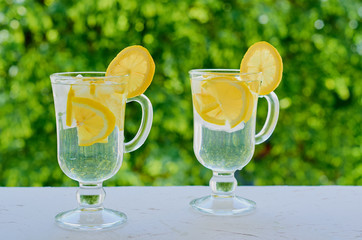 Lemonade water in the glasses on the blurred nature background. Cocktail with fresh lemon juice and ice. Healthy drinks, summer detox and diet concept