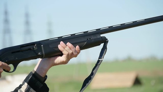 Profile of a man holding his single barrel rifle, targeting and shooting at a clay pigeon. A white cartidge flies away on a green range in slo-mo