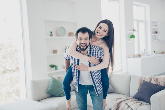 Portrait of playful foolish couple in casual outfits bearded man carrying his lover on back looking at camera in modern white apartment with interior