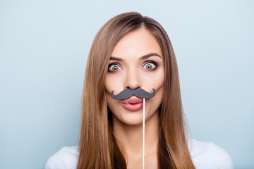 Close up portrait of funny foolish girl holding black carton mustache on stick with pout lips wide open eyes looking at camera isolated on grey background