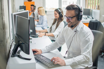 Obraz premium Smiling young man with headset using computer in call center.