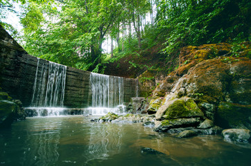 Beautiful green countryside. Waterfall on a river with big rocks and deciduous forest. Green landscape. 