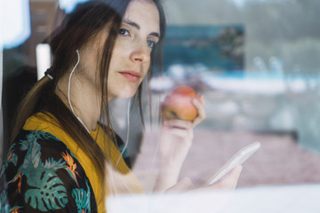 Young woman with earphones, cell phone and apple looking out of window