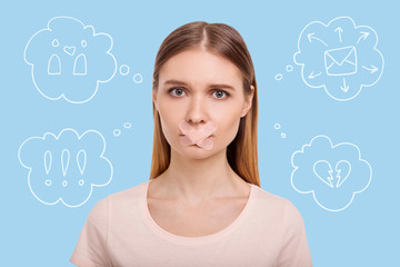 Unbearable silence. Portrait of young disappointed girl expressing sadness in eyes with a mouth being pasted with medical plaster while standing against blue background