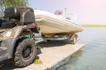 Obraz premium Transportation of inflatable boat on trailer. ATV quadbike moves ship to lake or river shore for launching. Beginning of water navigation and fishing season