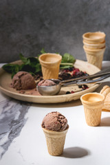 Homemade chocolate raspberry ice cream in small waffle cup served with frozen berries, fresh mint, chopped dark chocolate and metal spoon in ceramic plate on white marble kitchen table.