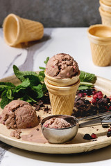 Homemade chocolate raspberry ice cream in small waffle cup served with frozen berries, fresh mint, chopped dark chocolate and metal spoon in ceramic plate on white marble kitchen table.