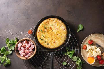 Spanish potato omelette tortilla with bacon served in cast-iron pan on cooling rack with ingredients above over over dark brown texture background. Top view, space.