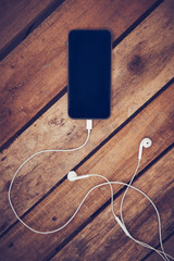 A top view of a set of wired earbud headphones and mobile phone blank template lying on a rustic wooden table. Styling and grain effect added to image.