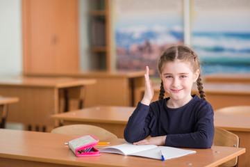 child girl sits at the Desk in the classroom, raises his hand, looks in the frame and smiling