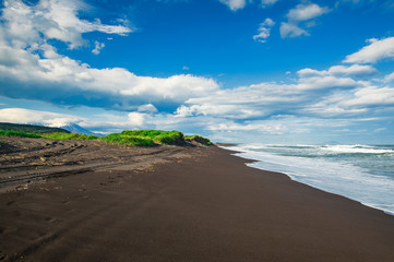 Halaktyr beach. Kamchatka. Russian federation. Dark almost black color sand beach of Pacific ocean. Stone mountains and yellow grass are on a background. Light blue sky