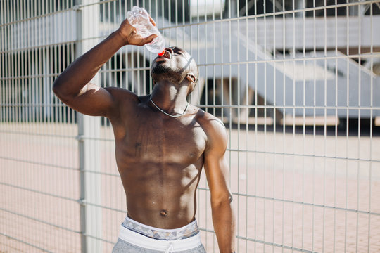 Handsome African American man with naked chest poses with a bottle of water before the fence in the morning
