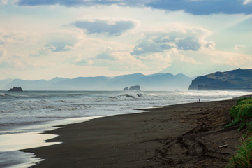 Fototapeta na wymiar Halaktyr beach. Kamchatka. Russian federation. Dark almost black color sand beach of Pacific ocean. Stone mountains and yellow grass are on a background. Light blue sky
