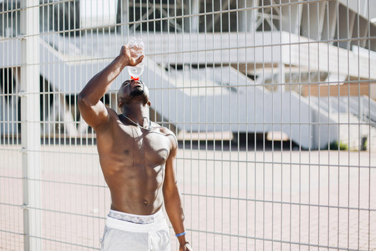 Handsome African American man with naked chest poses with a bottle of water before the fence in the morning