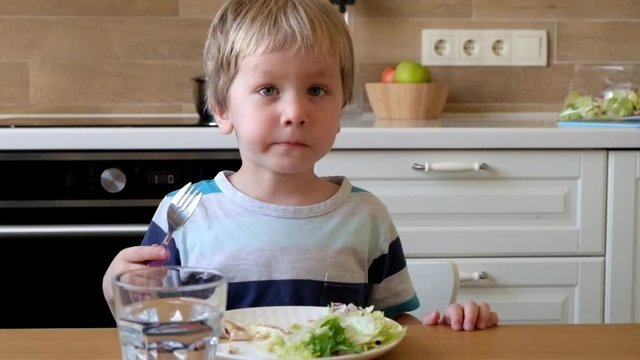 Little blond boy eating salad in the kitchen