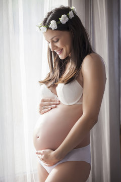 Pregnant happy woman in underwear by the window with curtains and a wreath of flowers on her head