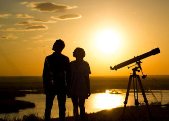 Silhouettes of couple - boy and girl near telescope on the hill at summer sunset