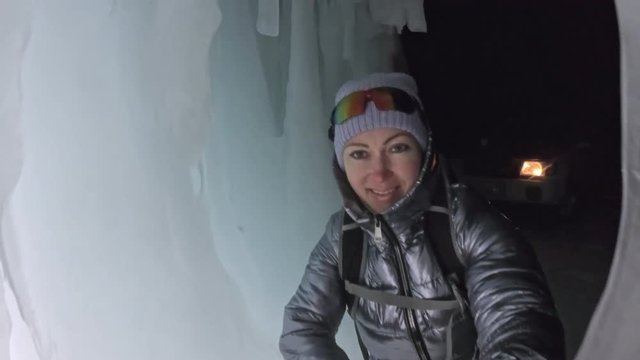 Travel woman on ice cave of Lake Baikal. Pov view selfie. Girl backpacker is walking of ice grot. Traveler looks at beautiful ice grotto. Hiker wears sports glasses, silver jacket, backpack, led lamp