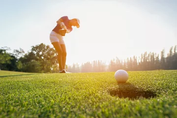 Papier Peint photo Golf Une femme asiatique de golfeur se concentre sur la balle de golf sur le golf vert le soir au coucher du soleil. Concept de mode de vie et de santé