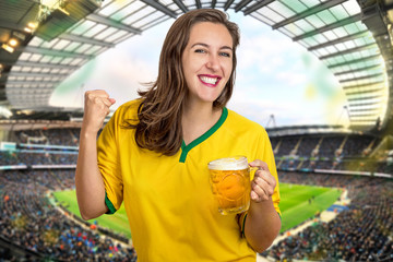 Girl in the stadium cheering for Brazil by drinking a very cold beer in the glass mug hoping that the Brazilian team will win the world cup along with the other fans.