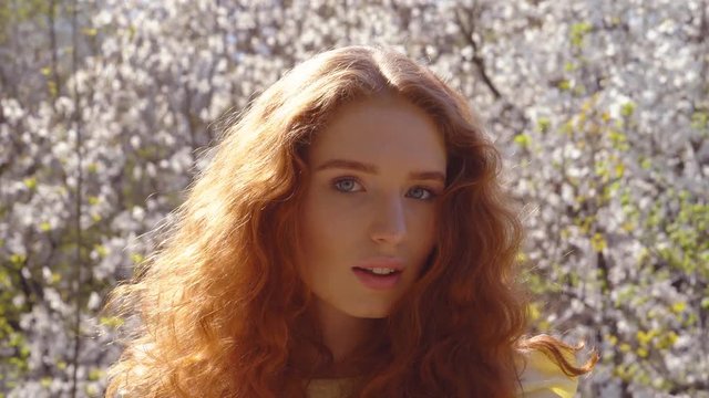 female portrait close-up. An amusing girl with natural red hair and blue eyes. in the background a blooming pink magnolia garden. spring freshness of the garden 4K