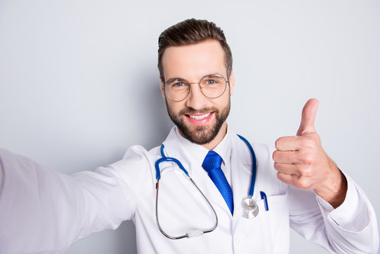 Self portrait of positive trendy doc in white outfit with tie and bristle having stethoscope on his neck shooting selfie showing thumb up, like sign with finger, hand over grey background