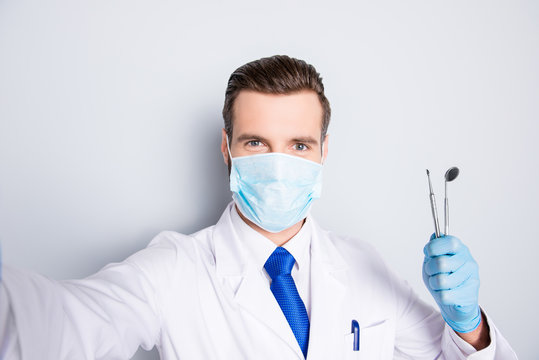 Self portrait of successful cheerful dentist in white lab coat, blue tie, shooting selfie on front camera, holding tools, having leisure, fun, video call, isolated on grey background