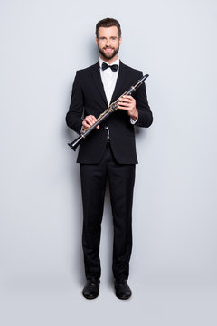 Full size, fullbody portrait of stylish, talented, cheerful musician with hairstyle in black tux with bowtie, pants, shoes holding bassoon in hands, looking ta camera, isolated on grey background