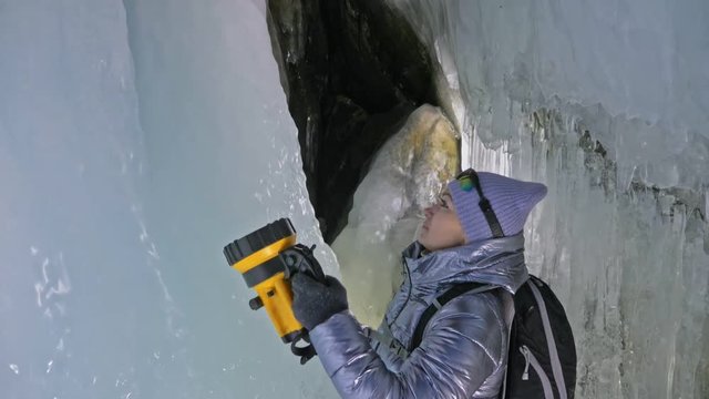 Travel woman on ice cave of Lake Baikal. Trip to winter island. Girl backpacker is walking of ice grot. Traveler looks at beautiful ice grotto. Hiker wears sports glasses, silver jacket, backpack, led