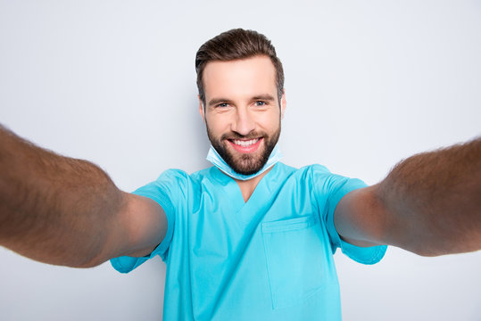 Self portrait of attractive cheerful man with stubble shooting selfie with two hands on front camera having sterile, protective, face mask, wearing lab blue uniform, isolated on grey background