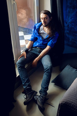 man with long hair and beard sits at home on a chair by the window