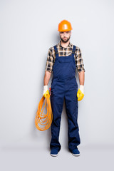 Full size fullbody portrait, snap of handsome strict electrician with bristle in overall, shirt, holding rolled cable in hand, looking at camera, isolated on grey background
