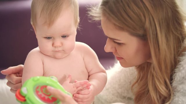 Mother playing with child. Close up of woman show green baby rattle to her daugther. Naked baby girl learning play. Sweet child sitting with mom. Infant playing with toy