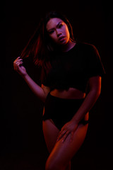 Fit young asian woman in black one piece poses against black under orange and blue colored lights