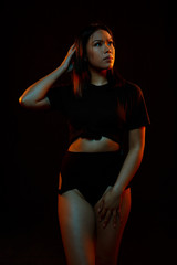 Obraz premium Fit young asian woman in black one piece poses against black under orange and blue colored lights