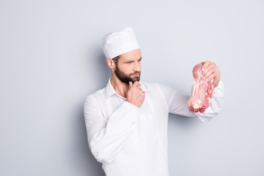 Portrait of pensive thoughtful butcher having fresh meat in hand looking at tenderloin holding hand on chin isolated on grey background
