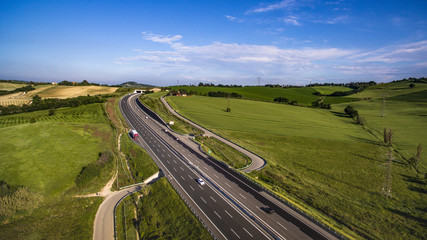 Italia, Maggio 2018 - Vista aerea dell'autostrada con automobili e camion che attraversa le colline con i campi di grano non ancora maturo