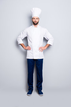 Full size fullbody portrait of attractive harsh chef cook with stubble in beret, holding arms on waist, isolated over grey background, looking at camera