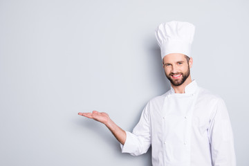 Portrait with empty place for advertisement, product of joyful attractive chef cook in beret with stubble holding copy space on his palm, looking at camera, isolated on grey background