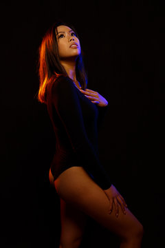Fit young asian woman in black one piece and necklace poses against black under orange and blue colored lights