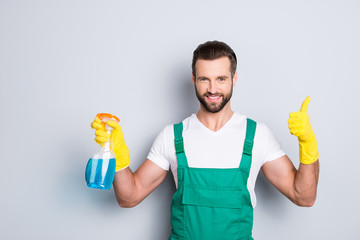 Portrait of cheerful joyful cleaner in uniform overall with stubble holding equipment in hand showing thumbup yes done recommend sign with finger isolated on grey background