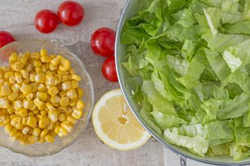 A colander with green salad. Background: cherry tomatoes, lemon and corn. From above.